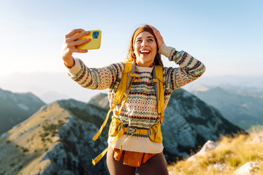 Hiker with bright hiking gear taking selfie on mountain. Cheerful woman feeling freedom and enjoying sunset on trail. Hiking, adventure concept. Active lifestyle.