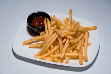 Golden crispy french fries served on a white rectangular plate with a small black bowl of ketchup sauce, isolated on light background