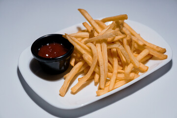 Golden crispy french fries served on a white rectangular plate with a small black bowl of ketchup dipping sauce, studio shot