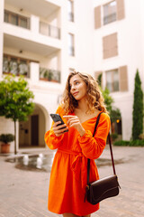 Fototapeta premium Beautiful woman in a stylish bright dress walks with her phone along a sunny embankment. A young tourist blogs, enjoys the weather on vacation. Travel concept, blogging.