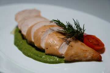Sliced grilled chicken fillet served on a white plate with green sauce and tomato, garnished with dill sprig in restaurant style