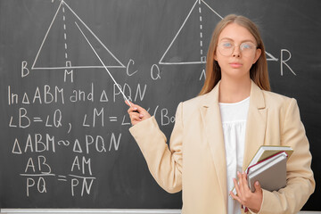 Young math teacher with pointer and books near blackboard in classroom