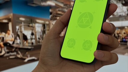 GREEN SCREEN CHROMA KEY A hand holding a phone with a green screen at a gym background filled with fitness equipment and active people exercising