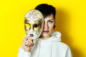 Beautiful young brunette woman holding carnival venetian mask on yellow background