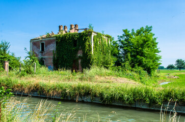 Un casale abbandonato e coperto dalla vegetazione lungo un canale di irrigazione nella campagna in una giornata estiva