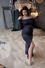 Fototapeta premium Beautiful pregnant woman in dark grey dress in sand barefoot. Big pregnant belly. Pretty brunette woman waiting child. Lady in nice room with lustres and portieres
