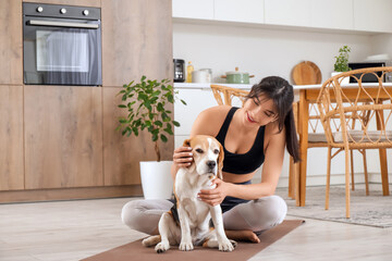 Pretty young woman playing with adorable Beagle dog on yoga mat in kitchen