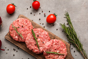 Cutting board with raw meat cutlets and rosemary on grey grunge background