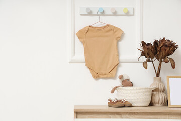 Commode with toy, dried flowers and baby bodysuit on light wall