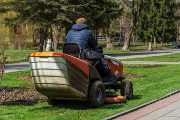 A man on a riding lawn mower trims the grass in a lush park surrounded by trees and scenic pathways on a bright spring day