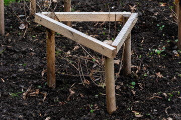 A triangular wooden frame stands on soil, supporting young plants as gardeners work in a spring garden, nurturing their growth amid fresh soil