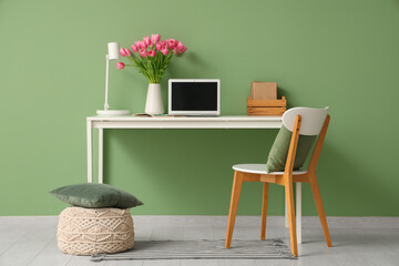 Stylish workspace with modern laptop, books and bouquet of beautiful tulip flowers near green wall