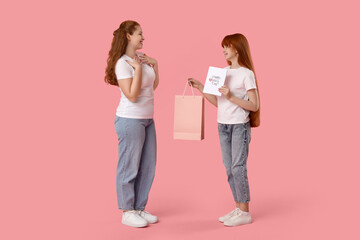Beautiful young woman and her cute daughter with shopping bag and greeting card on pink background....