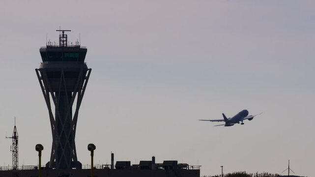 Large passenger aircraft ascending from airport runway, climbing swiftly past control tower silhouetted against dusky sky while gaining altitude and flying away toward distant horizon