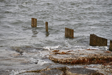 Old worn pier dock wood pillars in a stormy sea with waves breaking over rocks 