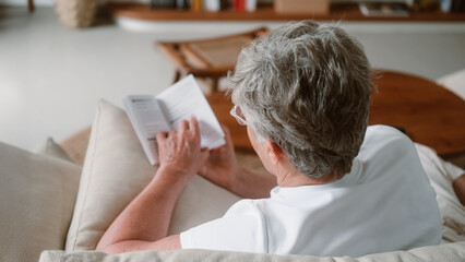 Naklejka premium A senior woman with white hair and glasses sits on a sofa in the living room and reads a paper book. Hobbies and leisure in retirement. Leisure and passion for fiction. Learning new information.