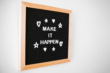 Phrase "Make It Happen" in white letters on a wooden and black felt board hanging on a white wall