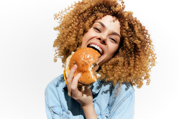 Smiling woman with curly hair holding a delicious hamburger, enjoying her meal against a bright...