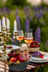 Two glasses of rose wine, flowers in a vase, silverware, fruits. Sunset, golden hour, atmospheric candlelight. Romantic table decor for a loving couple on blooming meadow with purple lupines