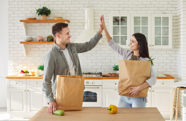 Cheerful young couple giving each other high five in kitchen while holding grocery bags. Happy man...
