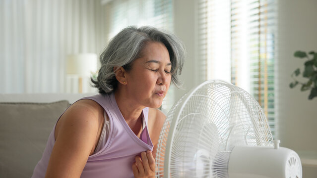 Asia adult people middle age old woman sit at sofa home feel hot flash fever use fan waving hand blowing wind face elder health care summer heat aging issue. Sad pain sweat tired stroke suffer stress.