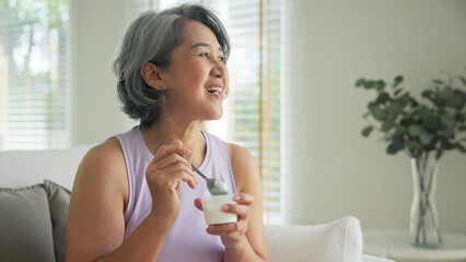 Asia adult people middle age happy old woman eat greek yogurt cup weight loss protein gut health care diet detox sit at home sofa elder aging asian aged female. Fresh medical food relief meal enjoy.