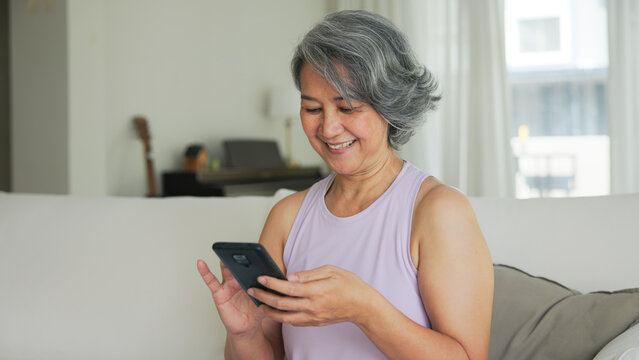 Asia adult people middle age happy relax old woman using mobile ask AI chatGPT chat bot look health care app sit at home sofa elder aging asian aged female. Online social media smart pay order system.