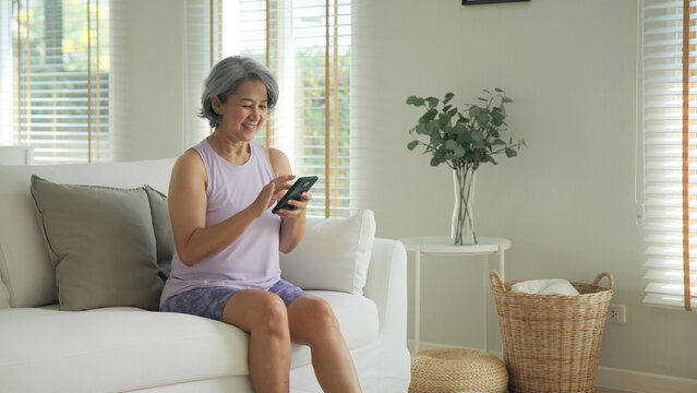 Asia adult people middle age happy relax old woman using mobile ask AI chatGPT chat bot look health care app sit at home sofa elder aging asian aged female. Online social media smart pay order system.