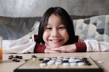 Portrait Close-up shot, one Asian girl fun playing Go, checkers board game on table in home living room, cheerful smile and looking at camera, happy weekend relaxing hobby of clever cute child winner.