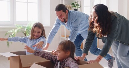 Happy family fun, parents playing with children in new modern cozy apartment living room, two cute little kids sitting in cardboard boxes, laughing mother and father pushing celebrating moving day