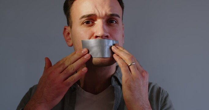 Close up portrait of young frustrated man covering his mouth with silver duct tape. Speechless guy staying silent, showing suppression of freedom. Human rights, freedom, truth, censorship concept.