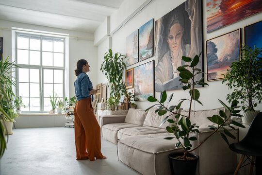 Female artist deeply reflects on portrait amidst other artworks on wall in studio. Creative workshop space with diverse collection of paintings, woman painter inspiration and new ideas for work