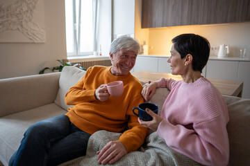 Obraz premium Joyful senior couple of husband and wife share laughter and stories, relaxing on comfortable couch with hot tea. Warm companionship, romantic hygge rest, glad smiling spouses during tea time at home