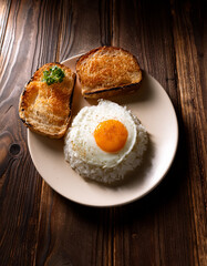 A slice of bread and a plate of rice topped with a sunny-side-up fried egg, both placed on a wooden table, natural lighting, high-resolution, food photography style