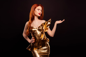 Elegant young woman in a golden dress with red hair and makeup posing against a dark background...