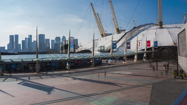 Timelapse of the O2 Arena's white dome and yellow supports, with pedestrians in the plaza and Canary Wharf's skyline under a sunny sky.