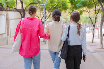Obraz premium Three young women walk together outdoors, seen from behind. They chat and enjoy each other's company under tree-lined streets. Concept of friendship, connection, and having fun in everyday life. 