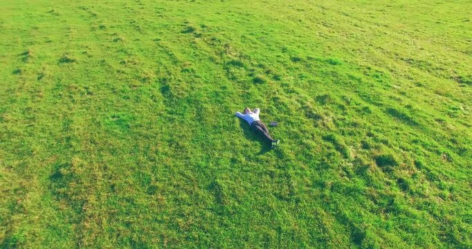 Aerial view point. Low orbital flight around man lied on green grass with notebook (pad) at yellow rural field. Casual weared. Sun rays. Radial movement.