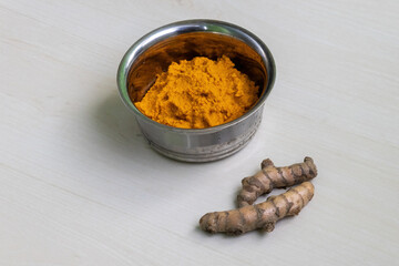 A steel bowl containing raw turmeric paste rests on a light wooden surface, alongside two whole raw turmeric roots.