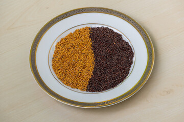 Black mustard and yellow mustard seeds on a white plate on a light wooden surface.