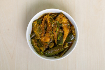 A delicious brinjal curry, cooked with hilsa fish (Tenualosa ilisha), is served in a white bowl on a light wooden surface. Top view.