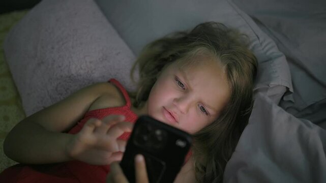 High angle: Young girl laying in bed, her face lit up by mobile phone