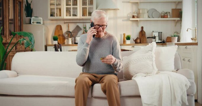 Lonely elderly man calling to call-center of bank, holding credit card in hands . Telephone scammers and financial fraud, security of personal data, aged male person talking by mobile phone at home
