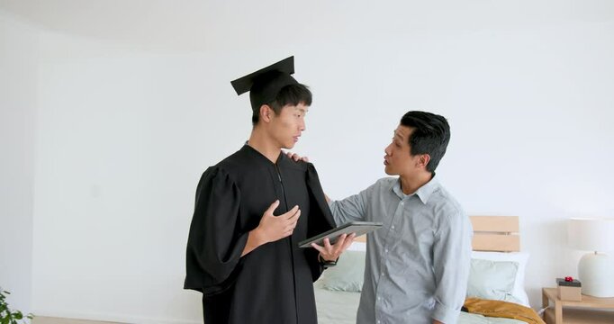 Father giving gift to son in graduation gown at home, celebrating achievement