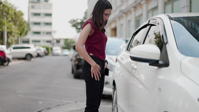 young asian woman walks to car and realizes key is missing, searches her pocket with concern, then turns back to pick it up from the ground on city street