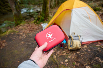 Closeup view of a hand holding a red first aid kit near a camping tent in the forest. First aid concept while traveling in the forest alone