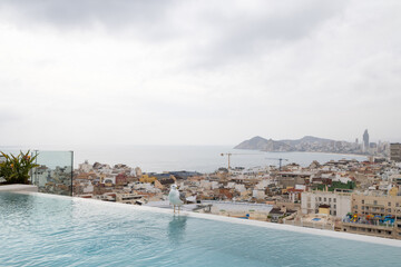 Photo of a white seagull bird sitting on the side of a swimming pool on a roof top with the Spanish town of Benidorm in the background on a hot but cloudy day