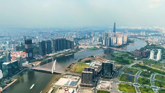 Panoramic aerial view of Ho Chi Minh City skyline with Saigon River, modern skyscrapers, and Landmark 81, Vietnam&rsquo;s tallest building, under a hazy blue sky.