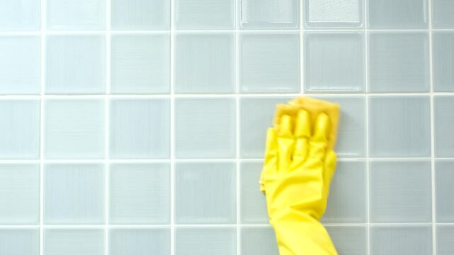 Female hand in yellow gloves wipes blue tiles in the bathroom, front view.