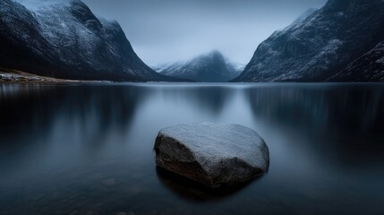 Calm lake, snow-capped mountains, serene winter landscape.  A lone rock sits amidst the still water reflecting the dramatic mountain range